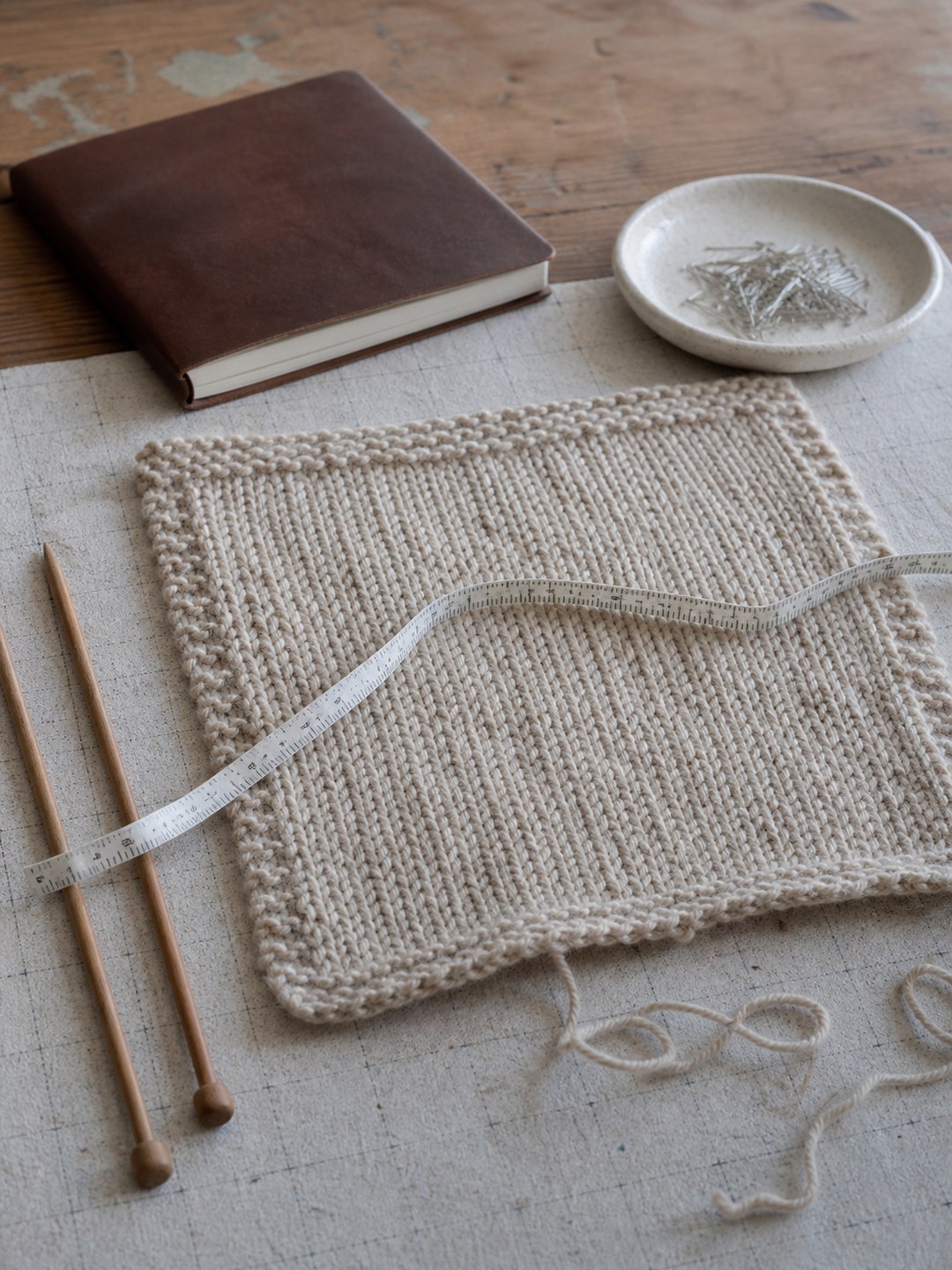 A washed oat-wool gauge swatch resting flat with wooden knitting needles, a soft measuring tape, loose yarn, and a small dish of pins on a worn worktable.